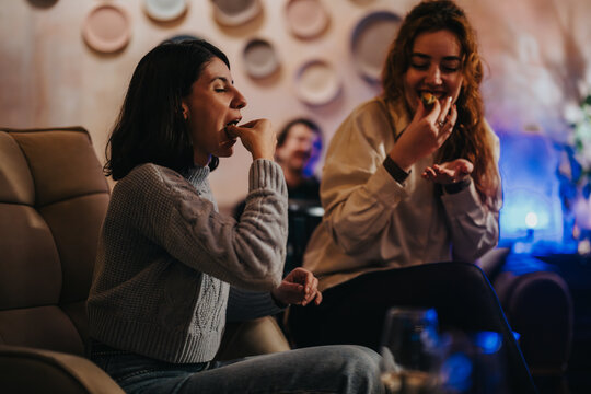 Two women are sharing a lighthearted moment while savoring snacks in a warmly lit lounge, embodying leisure and comfort.