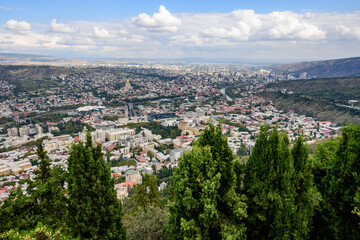 A sprawling aerial of Tbilisi, showcasing its mix of modern buildings and traditional architecture under a bright sky. 