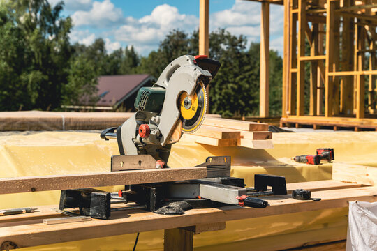 Circular saw resting on wooden plank at construction site with timber frame house being built