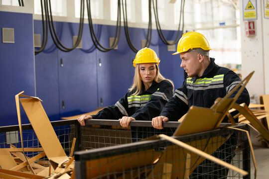 Male and female factory workers wearing safety gear checking cardboard waste in industrial bin. Team analyzing recycling process or material inspection in manufacturing site.