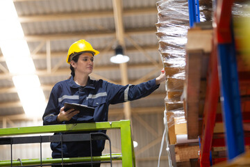 Female warehouse worker in safety gear inspecting pallet stock on high shelf using clipboard. Industrial logistics staff checking inventory in storage facility for supply chain management.