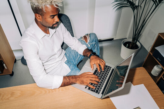 From above direct flash shot of Middle Eastern man in white shirt and ripped jeans typing and entering data into netbook while sitting near table and working in office.