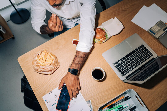 Direct flash top view of anonymous man in white shirt eating fries and burger and taking cellphone with black screen from desk near netbook while having lunch break in office.