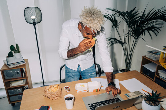 High angle direct flash shot of Middle Eastern male employee in smart casual clothes with blond dreadlocks biting burger and browsing data on netbook while standing near table and working in office.