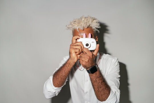 Unrecognizable young guy with dyed hair in white shirt taking photo on film camera against white background