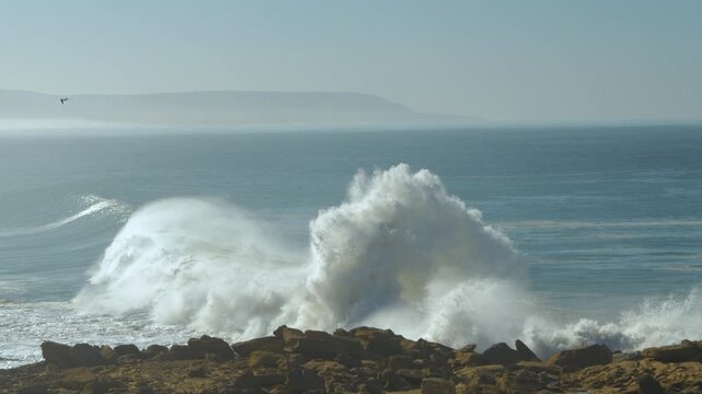 SLOW MOTION: Large cresting wave crashes against the rocks, raising a huge cloud of spray. Hazy horizon and distant coastline further emphasize the sense of the size and vastness of the wavy ocean.