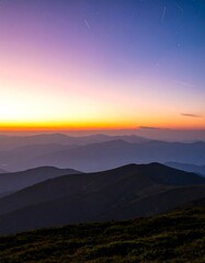Fototapeta premium Vibrant twilight sky over layered mountain peaks. Stunning sunset or sunrise colors illuminate the horizon above rolling hills. Serene natural landscape.