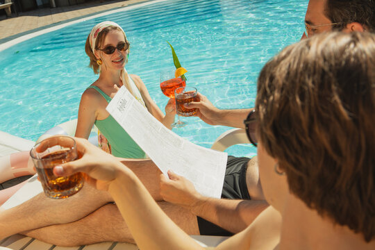 Friends relaxing by poolside with summer drinks and newspaper under bright sunlight. Concept of vacation lifestyle, summer leisure, tourism advertising, hospitality promotion.