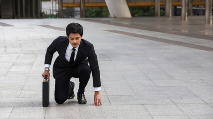 Southeast Asian businessman in suit crouching at starting line on urban pavement, holding briefcase...