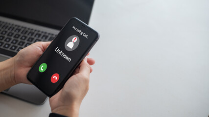 Close-up of hands holding smartphone showing incoming call from unknown number with warning icon, representing scam alert, spam call, privacy protection, and mobile communication security issues.