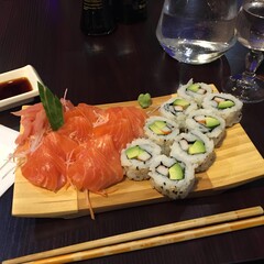 Elegant wooden sushi platter served at a Japanese restaurant: salmon sashimi, California rolls, soy sauce, wasabi and chopsticks arranged on a dark wood table.
