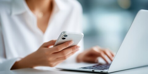Person using smartphone while working on laptop in modern office environment with soft natural light