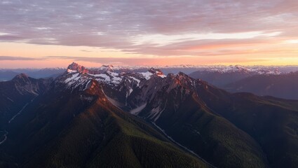 Naklejka premium Aerial view of snow capped mountains during sunset with a colorful sky and green forested valleys