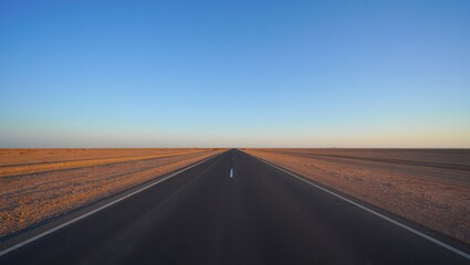 A long straight asphalt road stretches through a desert landscape under a clear blue sky at sunset