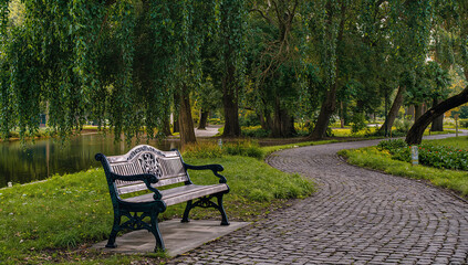 Fototapeta premium Lonely wooden bench beside quiet water in green park, cobblestone walkway under shade trees, peaceful outdoor nature scene 4k image