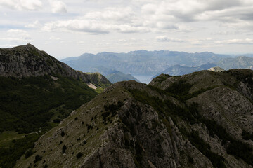 Fototapeta premium Scenic view from Mount Lovcen in Montenegro, featuring rugged mountain peaks, green valleys, and the distant Bay of Kotor. Dramatic clouds and layered mountain ranges create a breathtaking panoramic