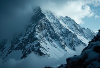 Obraz premium Majestic snow-covered mountain peak surrounded by clouds and mist under a partly cloudy sky