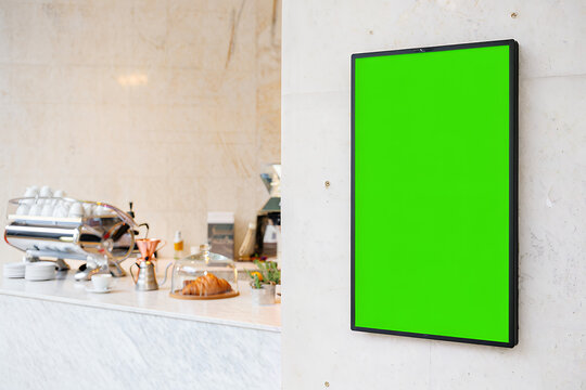 A vertical green screen digital menu board hangs on a wall in a modern, bright coffee shop interior.