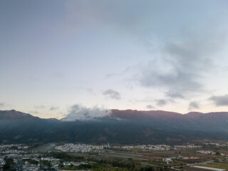 A panoramic view of a mountain range with a town nestled below, under a cloudy sky. The mountains are silhouetted against the horizon, with a hint of sunrise.