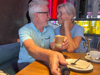 Joyful senior couple sharing a warm moment in a modern café. The man holds a selfie stick while the woman looks at him lovingly, capturing the essence of love, connection, and active senior lifestyle