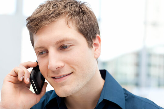 Man in dark blue collared shirt smiling while talking on smartphone in office with glass facade