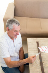 Fototapeta premium Senior man sitting holding cards in living room by coffee table with beige couch, copy space