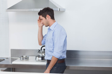 Man in twenties leaning against countertop in kitchen under range hood holding forehead, copy space