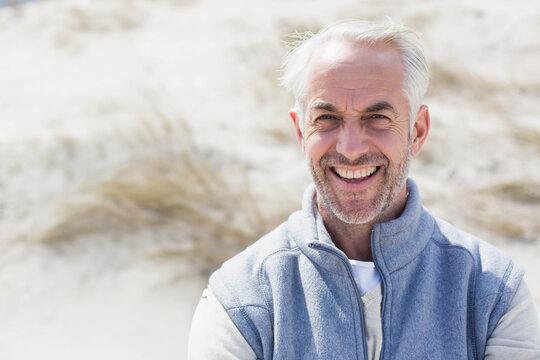 Senior man smiling and standing at coastal sand dunes wearing grey fleece vest, copy space