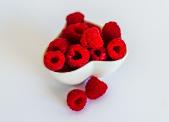 Small white heart-shaped ceramic bowl filled with bright red raspberries on a white surface. Some berries placed casually beside the bowl.