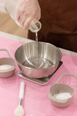 Cooking process, female hand in disposable transparent glove pouring water from carafe into steel bowl, food scales, pink dough mat, measuring spoons, brush