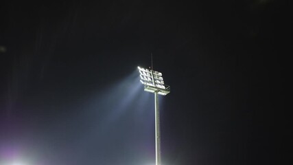 Bright stadium floodlight tower shining into the dark night sky with beams cutting through the air creating a dramatic and powerful sports atmosphere - Powered by Adobe