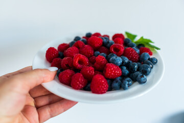 Hand holding a white plate filled with fresh raspberries and blueberries, garnished with mint. Shot in natural light on white background.