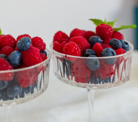 Two glass dessert bowls filled with fresh raspberries, blueberries, and mint leaves. Natural lighting and soft shadows on a white surface.
