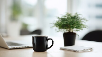 A black coffee mug sits on a wooden desk with a silver laptop and potted plant nearby,