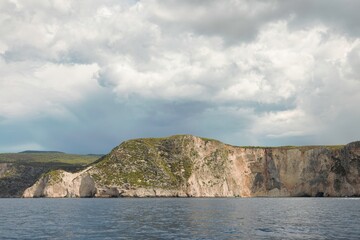 Fototapeta premium A coastal landscape in Greece shows a dramatic cliff face rising from the Ionian Sea under a sky filled with gathering storm clouds on a bright, overcast day
