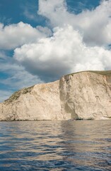 Naklejka premium A towering cliff face descends to the ocean, reflecting light in ripples. A vast cloudscape above dominates, offering a sense of scale in Zakynthos, Greece