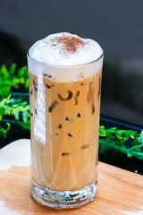 Glass of iced cappuccino topped with frothy milk foam and cocoa powder, placed on wooden surface with green leaves in background.