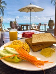 A low-angle shot captures a delicious breakfast served on a wooden table at a tropical beachside cafe. In the foreground, a white plate holds scrambled eggs, cucumber slices, shredded carrots, and two