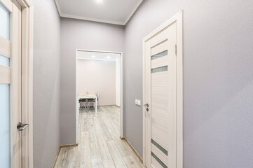hallway featuring light wood-patterned flooring, light gray walls, and doors with light wood frames. A glimpse into a room with a table and chairs can be seen through an open doorway