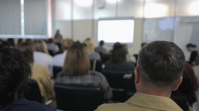 Man is sitting in a classroom with many other people. The man is looking at a projector screen