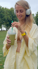 Woman Enjoying Green Smoothie and Music in Park by the Sea