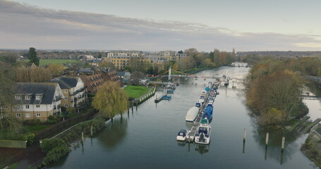 Fototapeta premium London, Teddington: Boats moored along the River Thames in Teddington, London city, create a serene scene with residential buildings houses and trees under a cloudy sky. Drone flight