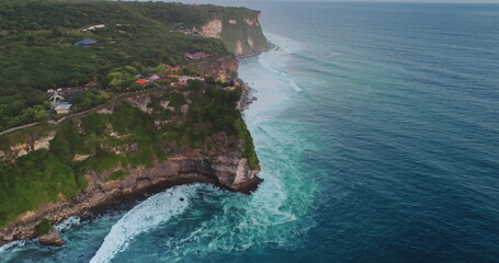Aerial drone view of Uluwatu Temple standing on top of a cliff during a cloudy day, waves crashing on rocks and lush vegetation covering the cliff in Bali, Indonesia
