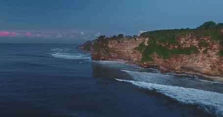 Soft pink and purple hues paint the sky as the sun sets over the dramatic cliffs of Pantai Dreamland Beach in Bali, Indonesia, with waves crashing against the rocky shore