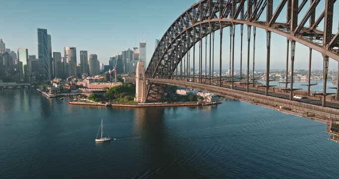 Australia, Sydney: Sydney Harbour Bridge and city skyline, sailboat glides under sunny morning blue sky. Travel destination, modern cityscape architecture in background. Aerial view drone