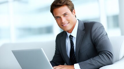 A confident young man in a suit smiles while working on a laptop, portraying professionalism in a modern office setting.