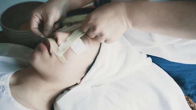 A woman is getting her face done by a man. The woman is wearing a white towel and the man is holding a white cloth over her face