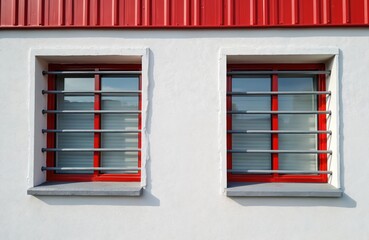 Obraz premium Two modern windows feature red frames, protected by gray security bars against textured white wall. Above, red corrugated metal roof adds contrasting industrial element. Scene emphasizes urban