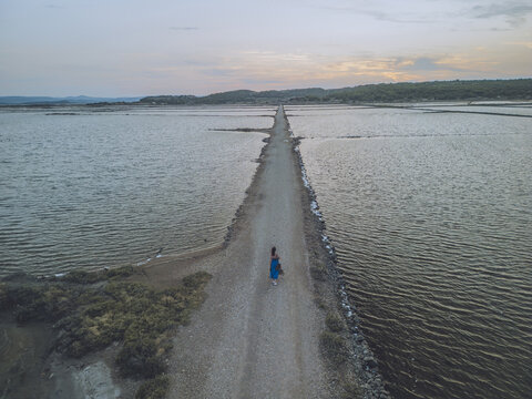 Aerial view of a lone figure walking along a narrow path cutting through the tranquil waters of a salt marsh, creating a striking contrast against the muted sky, Gruissan, Occitanie, France.