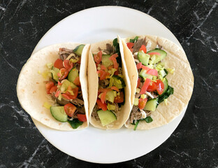 Three meat tacos on a white plate on a black marble worktop, with pulled meat, cucumber, tomato, lettuce and pickled radish for a healthy vibrant meal or snack